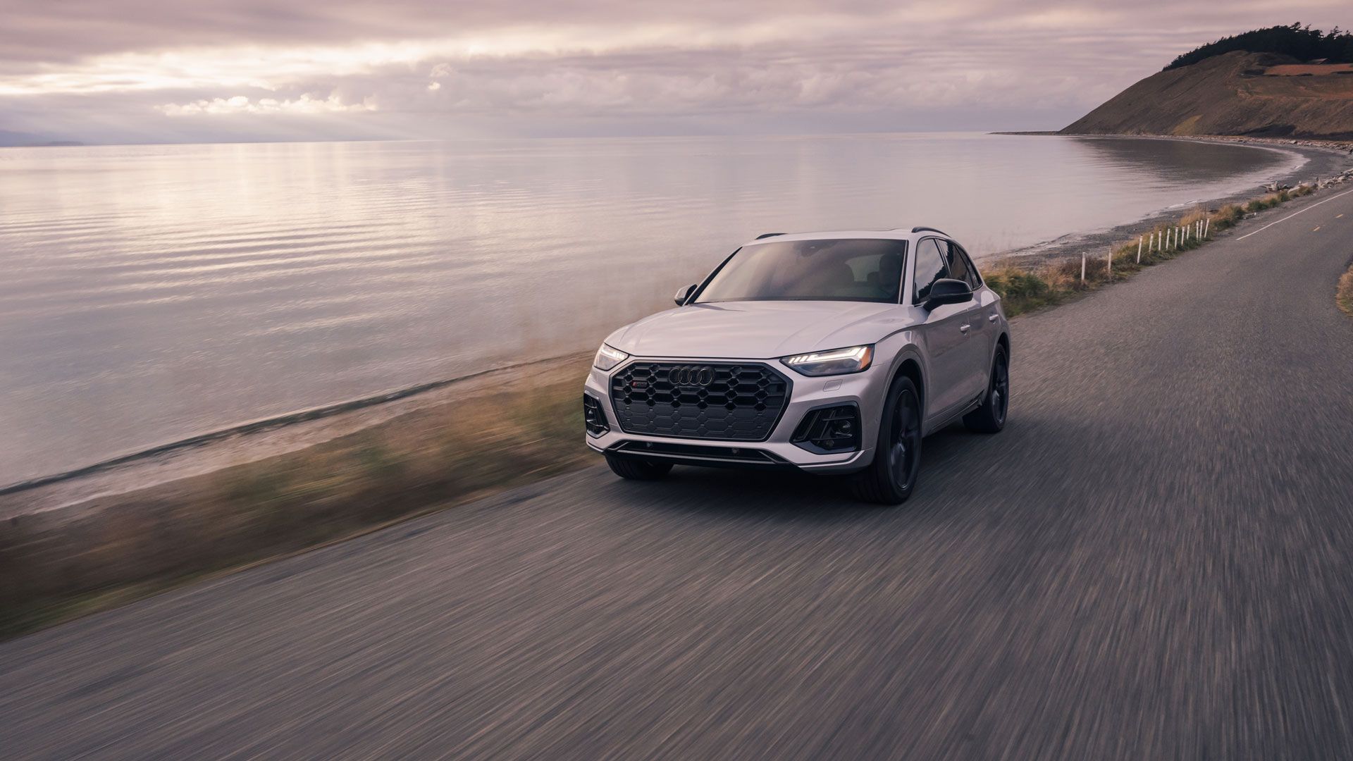 Light gray 2025 Audi Q5 on coastal road with ocean and mountains in the background.